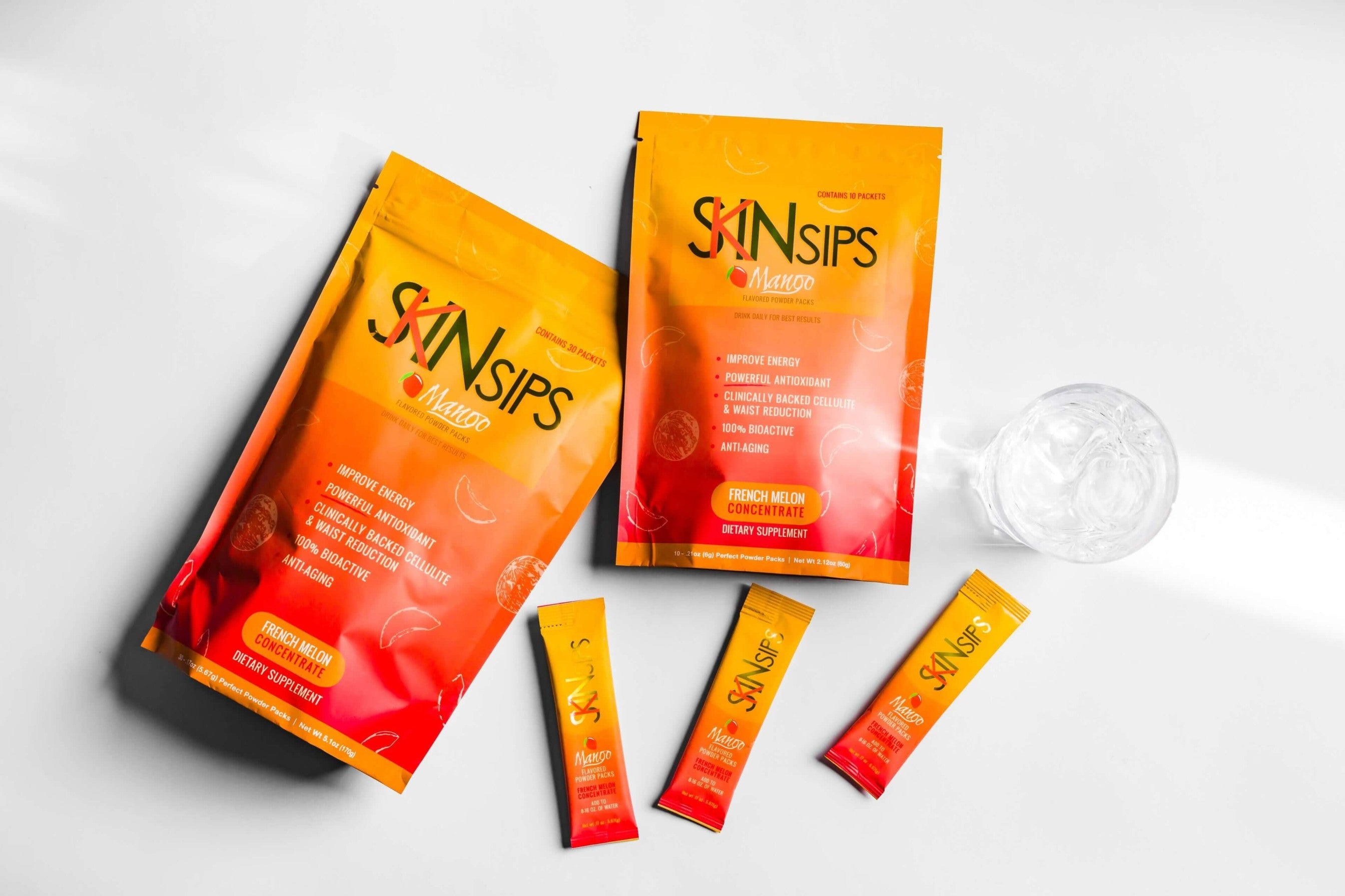 Two orange and yellow SkinSips Mango supplement bags with three single-serve powder sticks arranged beside a glass of water on a white background.