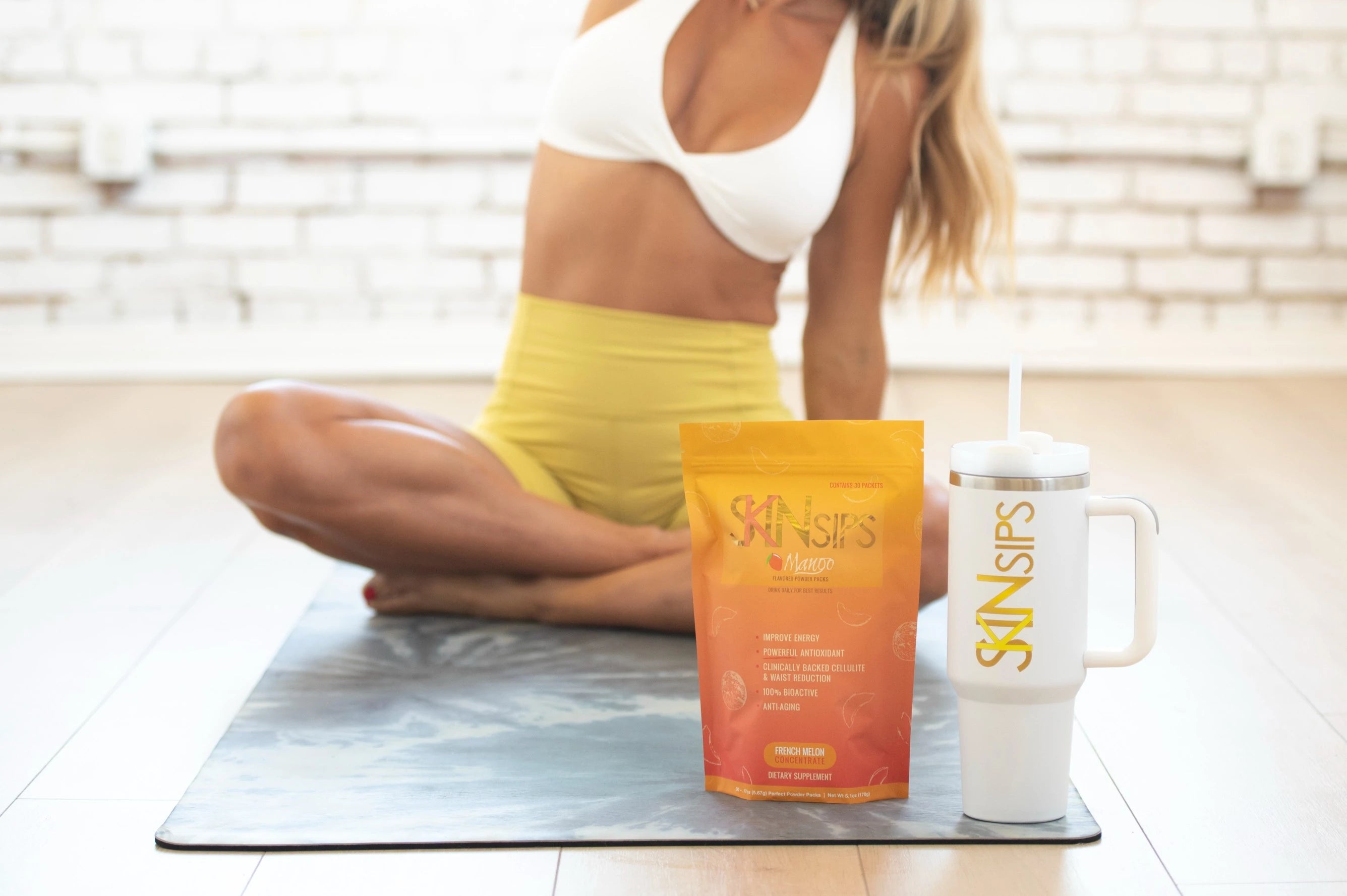 SkinSips mango pouch and tumbler on yoga mat beside woman in workout clothes.