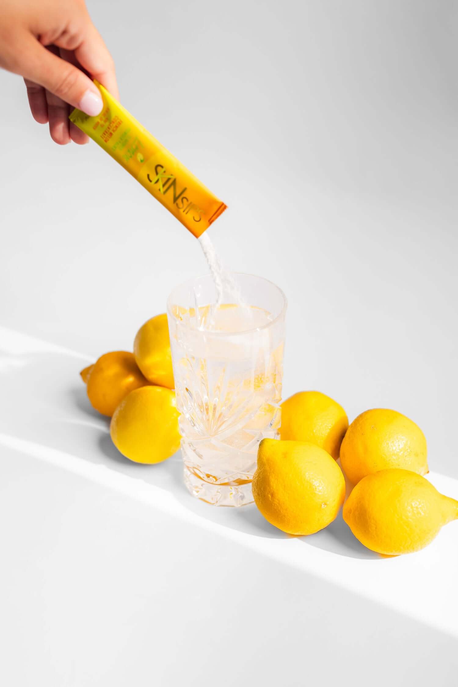 Hand pouring SkinSips lemon-flavored antioxidant powder into a glass of water, surrounded by fresh lemons.