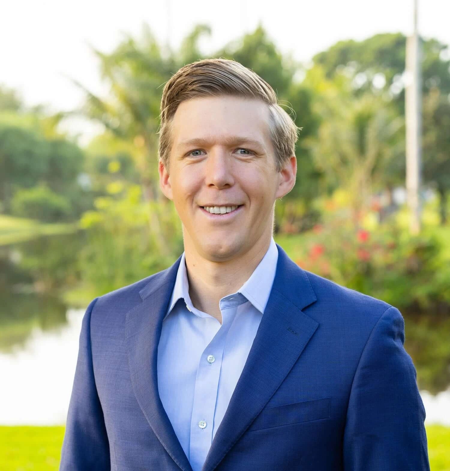 Dr. Leland Stillman in blue suit standing outdoors with greenery and water background.