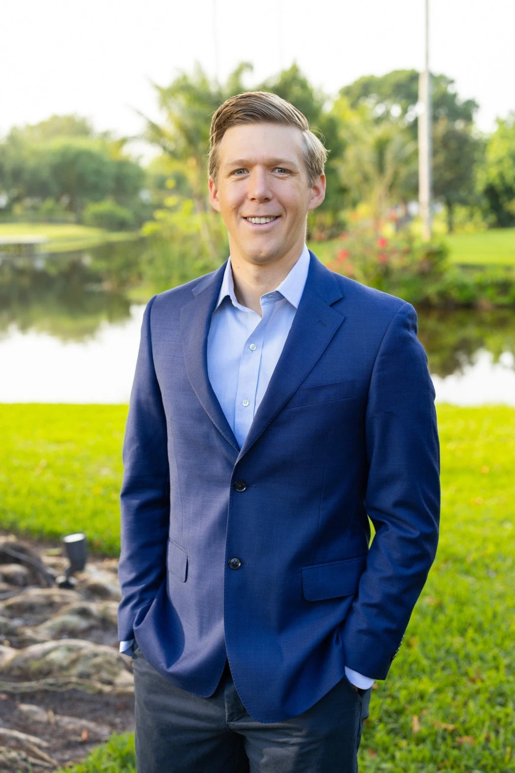 Dr. Leland Stillman in blue suit with hands in pockets standing outside by water and trees.