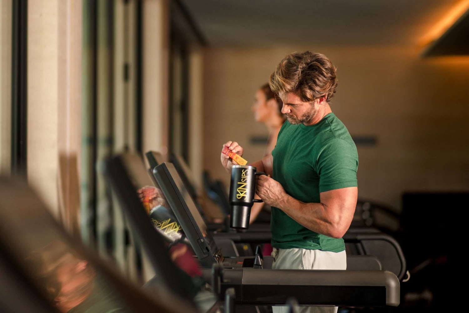 Man adding SkinSips Plus electrolyte powder to a drink bottle while walking on a treadmill at the gym.