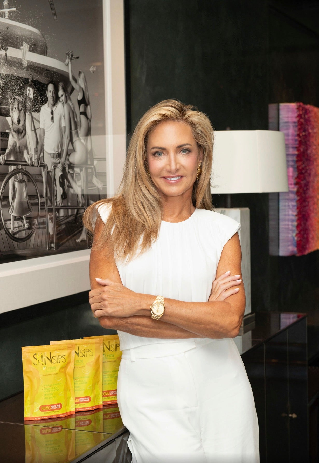 Mary Rankin standing confidently in a white outfit with arms crossed, smiling beside SkinSips product packets on a counter.