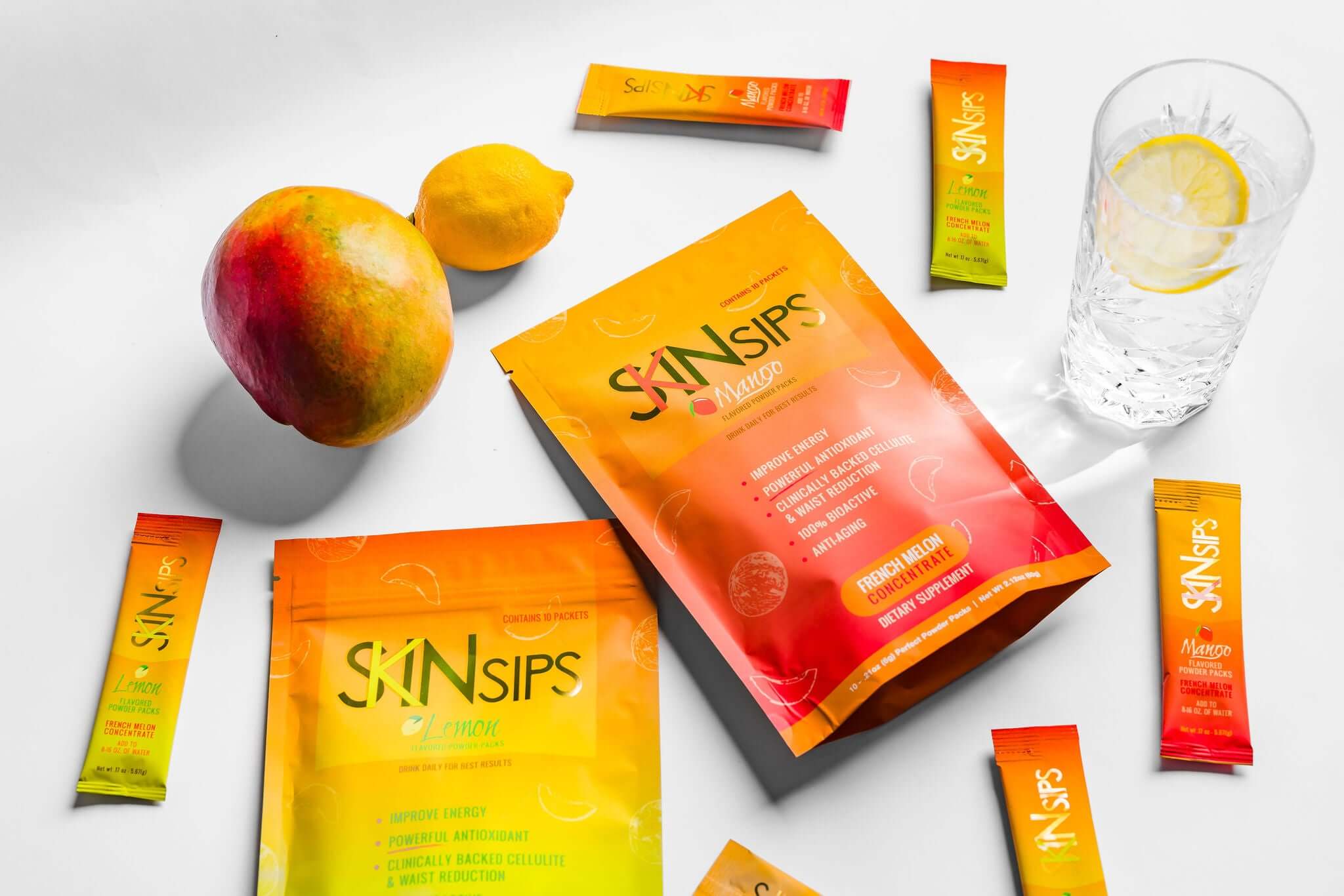 Flat lay of SkinSips Mango and Lemon supplement packets surrounded by single-serving sticks, a mango, a lemon, and a glass of lemon water.