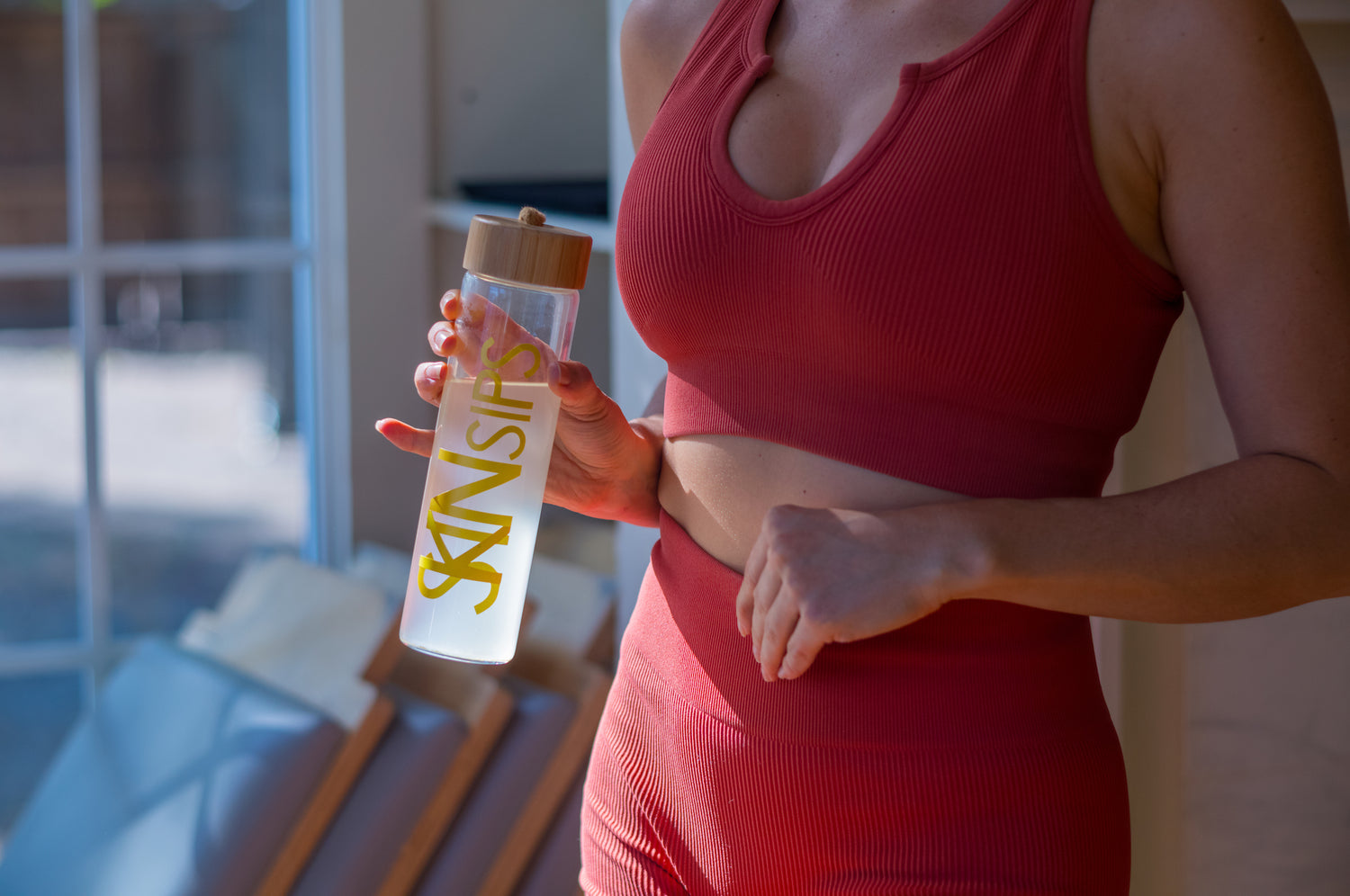 Woman wearing red workout attire holding a glass SkinSips branded watter bottle.