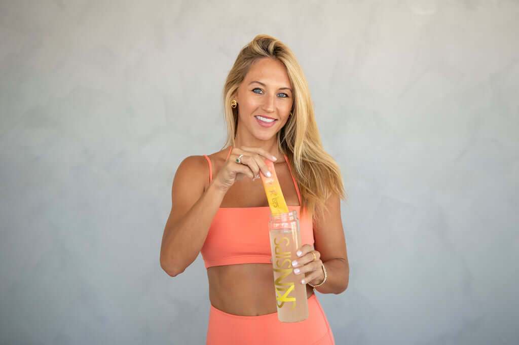 Smiling woman in a coral sports outfit holding a SkinSips drink mix stick above a clear shaker bottle filled with water."