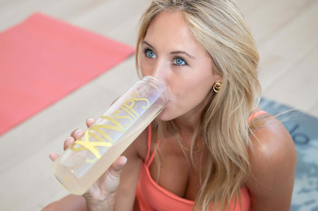 Close-up of a woman with blonde hair and blue eyes drinking from a clear SkinSips branded water bottle while sitting on a yoga mat.