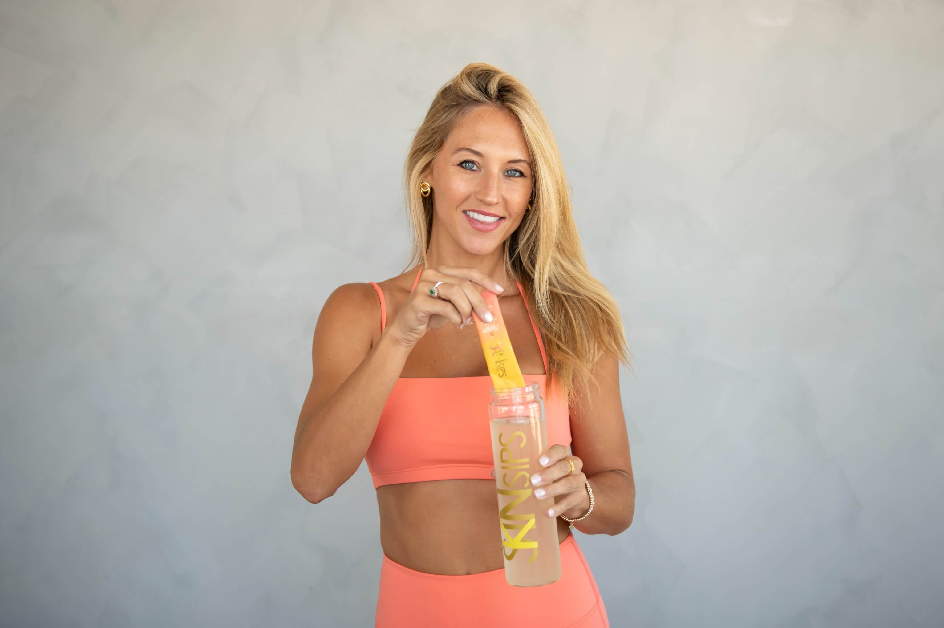 Smiling woman in peach workout set pours SkinSips powder stick into branded bottle."