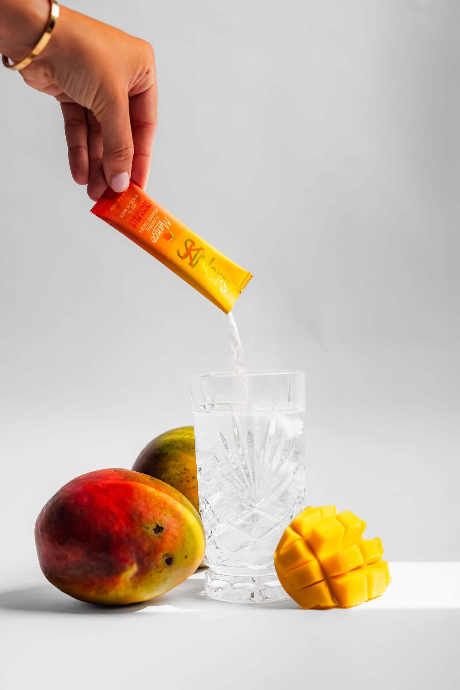 Hand pouring SkinSips Mango powdered supplement into a glass of water, with fresh whole and sliced mangoes arranged beside the glass.