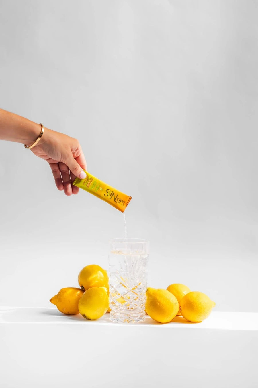 Hand pours SkinSips powder into glass of water surrounded by fresh lemons.